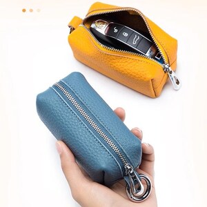 May include: Two leather key cases. One is open, revealing a car key inside, and is a bright yellow-orange color. The other is a blue-grey color and is closed, with a silver zipper and a silver clasp.