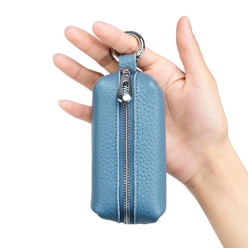 May include: A blue leather key case with a silver zipper and key ring. The key case is held in a hand, showcasing its compact, rectangular design. The zipper runs vertically down the center, providing easy access to the keys.