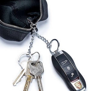 May include: Black leather key pouch with a silver chain and keyrings. Attached are several keys and a black car key fob with the Porsche logo.