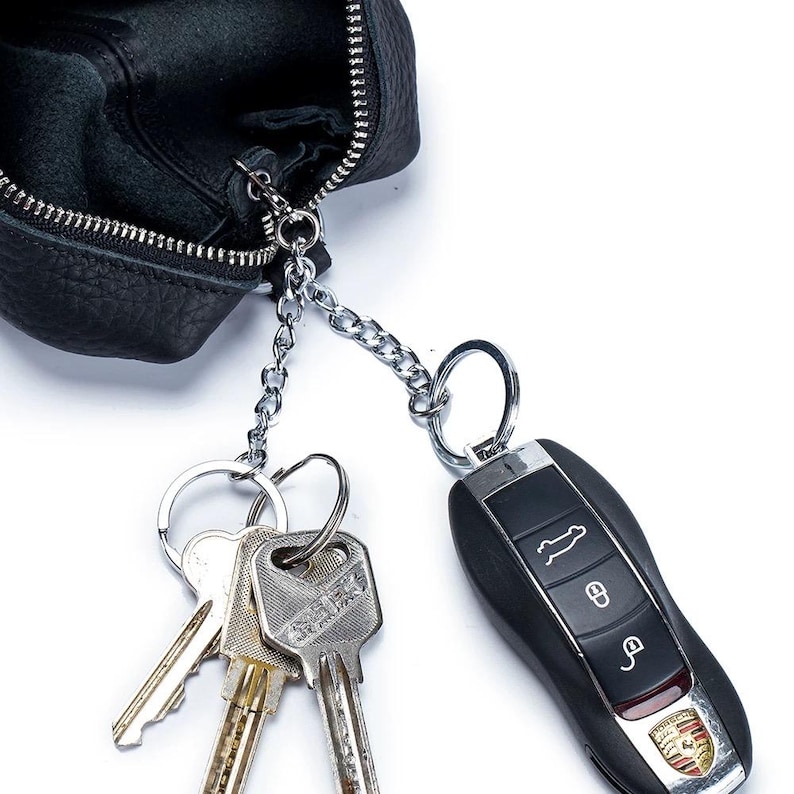 May include: Black leather key pouch with a silver chain and keyrings. Attached are several keys and a black car key fob with the Porsche logo.