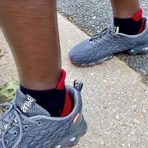 May include: Gray athletic shoes with orange soles and gray laces. The shoes are paired with black socks with red and green accents. The shoes are on a concrete surface.