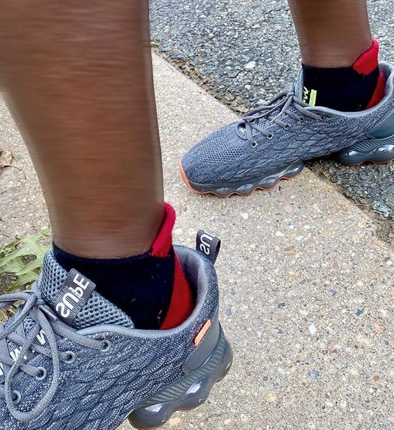 May include: Gray athletic shoes with orange soles and gray laces. The shoes are paired with black socks with red and green accents. The shoes are on a concrete surface.