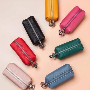 May include: An assortment of colorful key cases in various shades, including yellow, pink, green, blue, red, black, and light pink. Each case is rectangular with a zipper closure and a silver-toned key ring attachment. The cases are made of a textured material.