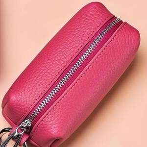 May include: A vibrant pink leather key case with a silver zipper closure and a silver key ring. The case has a textured surface and a rectangular shape, perfect for organizing keys. The background is a soft peach color.