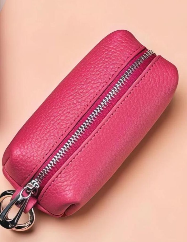 May include: A vibrant pink leather key case with a silver zipper closure and a silver key ring. The case has a textured surface and a rectangular shape, perfect for organizing keys. The background is a soft peach color.