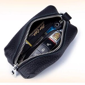 May include: Black leather key case with a silver zipper, open to reveal a car key and a lipstick tube. The lipstick tube has the text "Keep Color" printed on it. The key case is rectangular and has a small loop on the side.