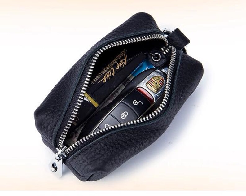 May include: Black leather key case with a silver zipper, open to reveal a car key and a lipstick tube. The lipstick tube has the text "Keep Color" printed on it. The key case is rectangular and has a small loop on the side.