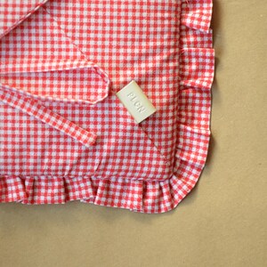 May include: A red and white gingham fabric item featuring a ruffled edge and a small rectangular beige tag with the embossed text PLCN. The textile has a classic checkered pattern and includes a corner section with a visible fabric tie.