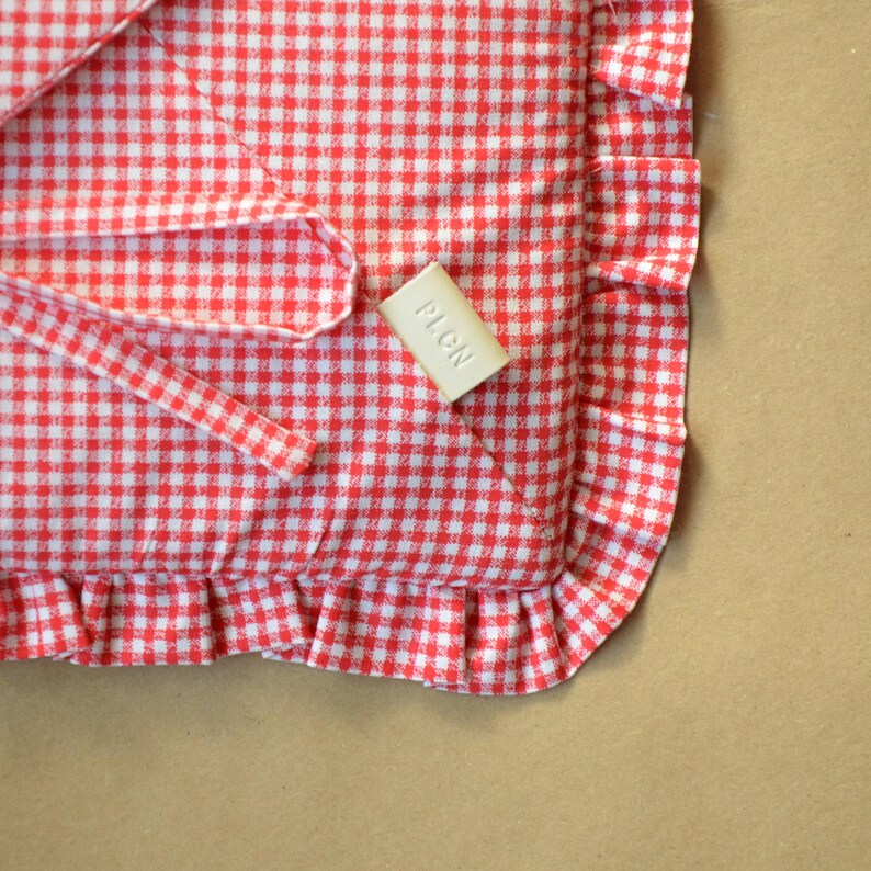 May include: A red and white gingham fabric item featuring a ruffled edge and a small rectangular beige tag with the embossed text PLCN. The textile has a classic checkered pattern and includes a corner section with a visible fabric tie.