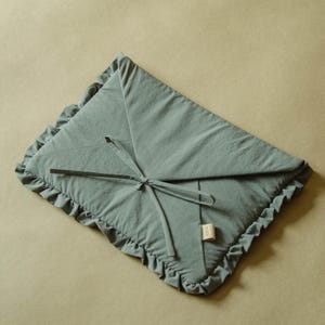 May include: A sage green rectangular fabric envelope case featuring a ruffled border along the edges and a tie closure made of matching fabric strings. A small rectangular beige fabric tag is attached to the side of the envelope case.
