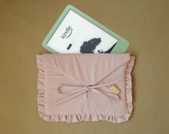 May include: A light green e-reader with a white screen displaying the Kindle logo and a black tree graphic, partially inserted into a dusty pink fabric sleeve featuring a ruffled border, a triangular envelope-style flap, and a tied ribbon closure.