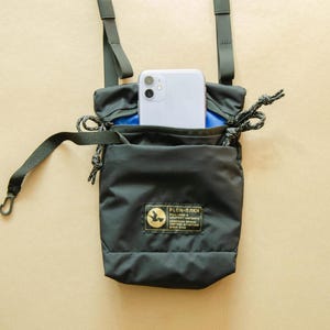 May include: A black crossbody bag with a blue interior and a small phone pocket. The bag has a black adjustable strap and a small black label with the text "PLCN-PLAK" on the front.