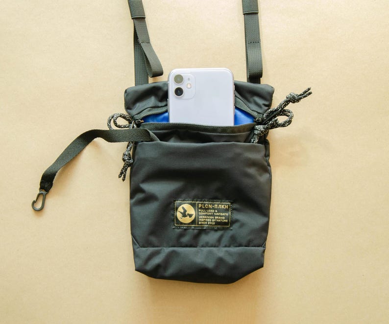 May include: A black crossbody bag with a blue interior and a small phone pocket. The bag has a black adjustable strap and a small black label with the text "PLCN-PLAK" on the front.