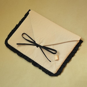 May include: A beige envelope-style pouch featuring a black ruffled trim along the edges. The pouch is secured with a black fabric tie closure tied in a bow. A small rectangular beige tag is attached to the lower right side of the pouch.