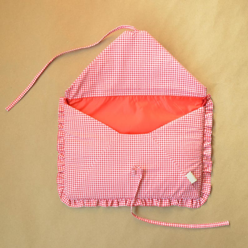 May include: A fabric envelope-style pouch featuring a red and white gingham check pattern. The pouch has a ruffled border, a solid red interior lining, and two long fabric ties attached to the top flap and the bottom edge for closure.