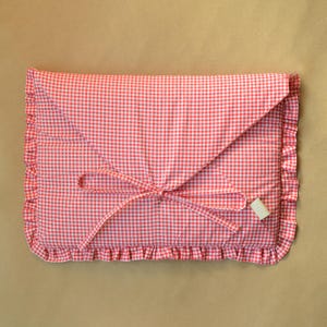 May include: A rectangular fabric pouch featuring a red and white gingham pattern. The envelope-style flap is secured with two thin fabric ties. The edges of the pouch are finished with a gathered ruffle trim, and a small rectangular tag is attached.