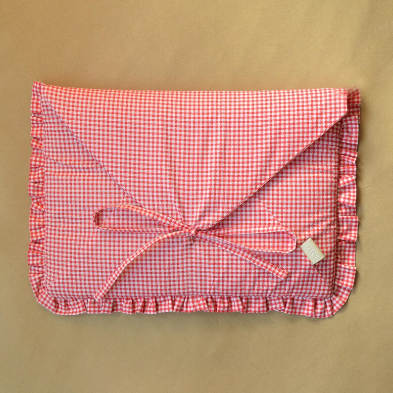 May include: A rectangular fabric pouch featuring a red and white gingham pattern. The envelope-style flap is secured with two thin fabric ties. The edges of the pouch are finished with a gathered ruffle trim, and a small rectangular tag is attached.
