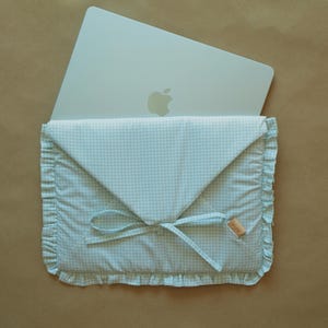 May include: A light blue and white gingham fabric laptop sleeve with a ruffled edge and a tie closure. The envelope-style case features a small tan rectangular tag with the text "FLOU" and holds a silver laptop with an Apple logo visible.