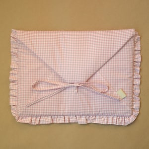 May include: A rectangular fabric envelope case featuring a light pink and white gingham pattern. The case has a triangular flap, a ruffled border along the edges, and a thin fabric tie closure. A small rectangular label is attached to the front.