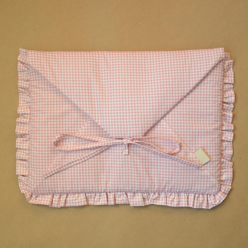 May include: A rectangular fabric envelope case featuring a light pink and white gingham pattern. The case has a triangular flap, a ruffled border along the edges, and a thin fabric tie closure. A small rectangular label is attached to the front.