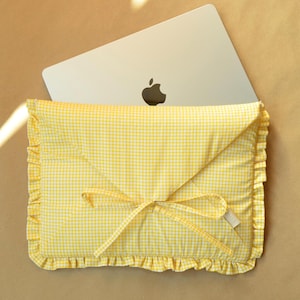 May include: A yellow gingham laptop sleeve with a ruffled edge and a bow closure. The sleeve is made of a soft, quilted fabric and is designed to protect a laptop. A silver laptop with the Apple logo is partially visible.