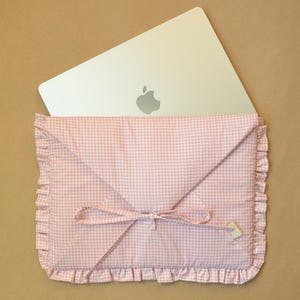 May include: A light pink and white gingham laptop sleeve with a ruffled border and a tie closure. The envelope-style sleeve features a triangular flap and is shown with a silver laptop partially inserted, displaying a visible Apple logo on the lid.
