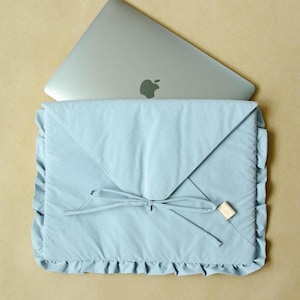 Könnte beinhalten: Eine hellblaue Laptoptasche mit Rüschenkante und Schleifenverschluss. Ein silberner Laptop mit dem Apple-Logo ist teilweise sichtbar. Die Hülle ist aus weichem Stoff und hat ein kleines Etikett.