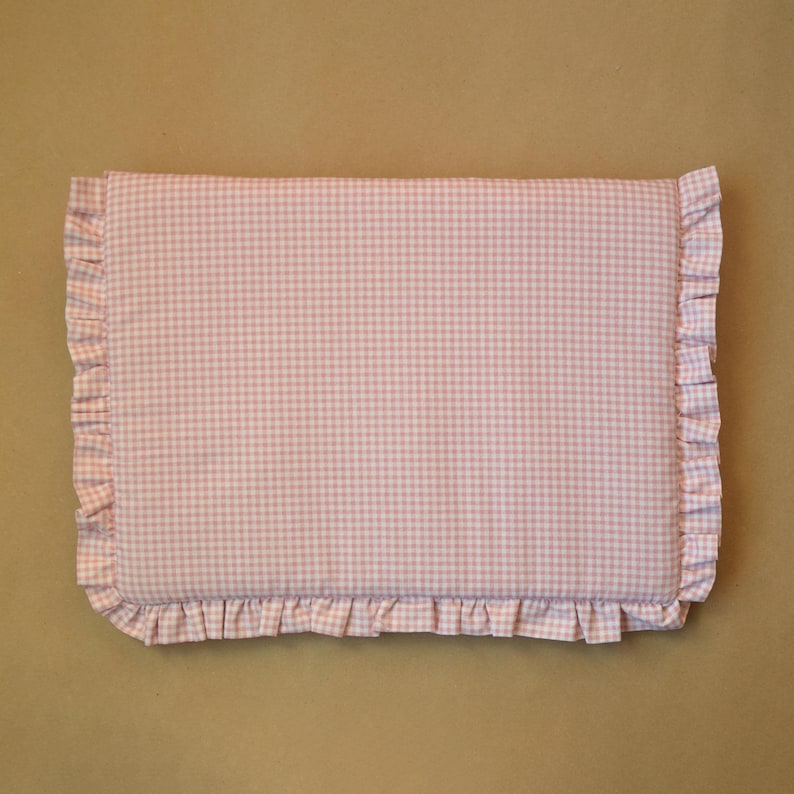 May include: A rectangular fabric pouch featuring a light pink and white gingham check pattern. The pouch is finished with a gathered ruffle trim along all four edges, creating a soft, textured border around the main checkered rectangular body.
