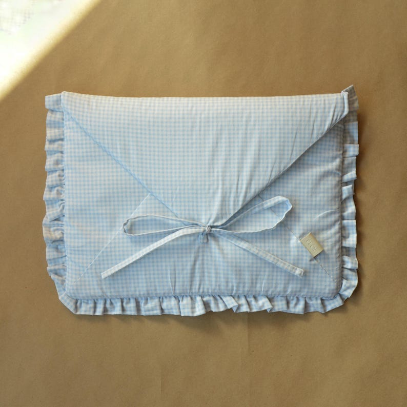 May include: A rectangular fabric envelope case featuring a light blue and white gingham check pattern. The case has a triangular flap closure secured with thin fabric ties and is finished with a ruffled border along the edges. A small tag is attached.