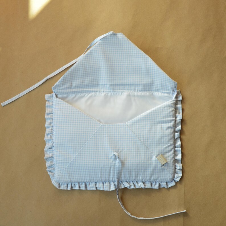 May include: A light blue and white gingham fabric envelope-style pouch featuring a ruffled border along the edges. The interior is lined with a solid white fabric, and the exterior includes a matching fabric tie closure attached to the flap.