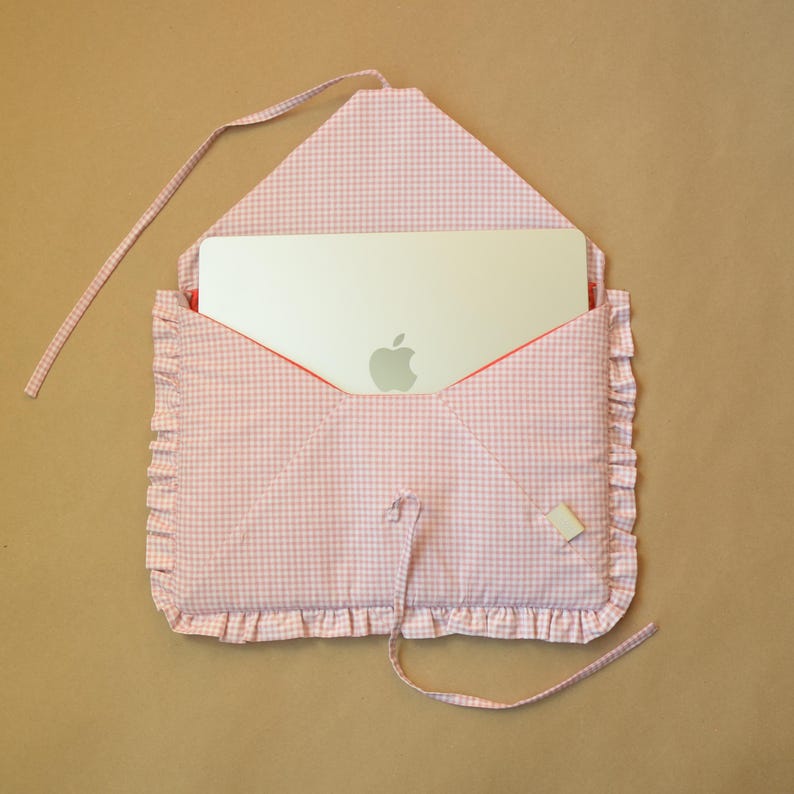 May include: A light pink and white gingham fabric laptop sleeve shaped like an envelope with a ruffled border. The sleeve features a tie closure and contains a silver laptop with an Apple logo visible on the back, partially tucked inside the case.