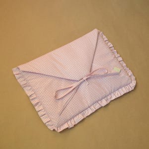 May include: A rectangular fabric pouch featuring a light pink and white gingham pattern. The pouch has a triangular flap closure secured with two thin fabric ties and is finished with a ruffled border along the entire outer edge of the item.