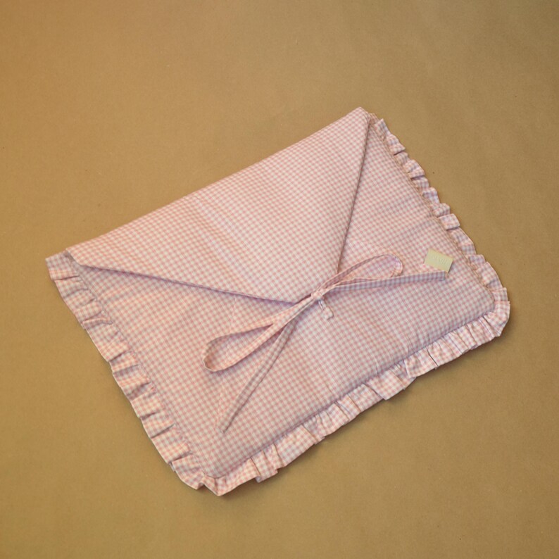 May include: A rectangular fabric pouch featuring a light pink and white gingham pattern. The pouch has a triangular flap closure secured with two thin fabric ties and is finished with a ruffled border along the entire outer edge of the item.