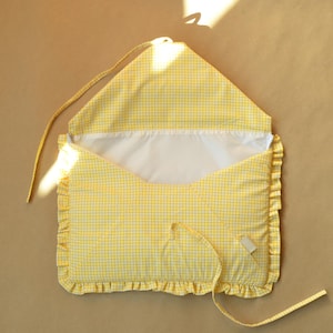 May include: A yellow and white gingham fabric envelope-style pouch featuring a ruffled border along the sides and bottom. The interior is lined with solid white fabric, and the flap includes a long fabric tie for closure and securing the contents.