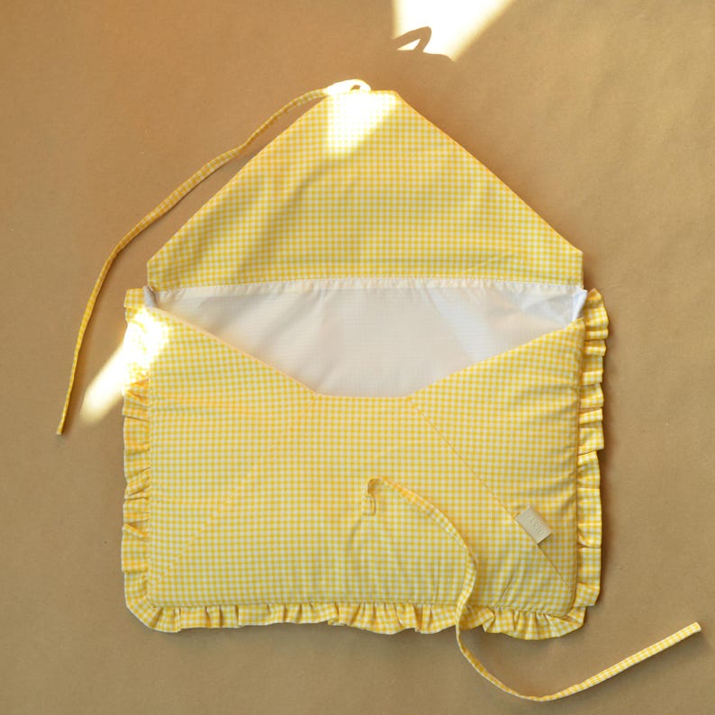 May include: A yellow and white gingham fabric envelope-style pouch featuring a ruffled border along the sides and bottom. The interior is lined with solid white fabric, and the flap includes a long fabric tie for closure and securing the contents.