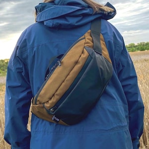 Puede incluir: Un bolso bandolera marrón y negro con un compartimento principal con cremallera y una correa ajustable. El bolso presenta una pequeña etiqueta rectangular con el texto PLCN en la esquina inferior frontal, llevado en diagonal sobre la espalda sobre una chaqueta azul.