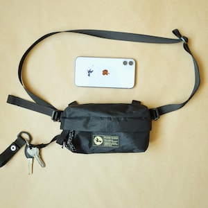 Puede incluir: Un bolso bandolera negro con correa ajustable, que presenta un parche rectangular con un logotipo de pájaro amarillo y el texto PLCN-PLKN. Un teléfono inteligente plateado se coloca sobre el bolso y dos llaves de metal cuelgan de una correa.
