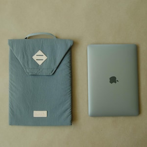 Op de afbeelding: Een teal laptophoes met een handvat en een zilveren laptop. De hoes heeft een klepsluiting met een ruitvormige patch en een klein rechthoekig label. De laptop heeft het Apple-logo.