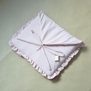 May include: A pale pink baby blanket with a frilled edge and a tie fastening. The blanket is made of soft material and has a label attached. The blanket is folded into a square shape.