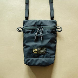 May include: A black crossbody bag with a zipper closure and a small patch with the text "PLCN-ПЛАЙК" and a bird logo. The bag has a black adjustable strap and a drawstring closure.