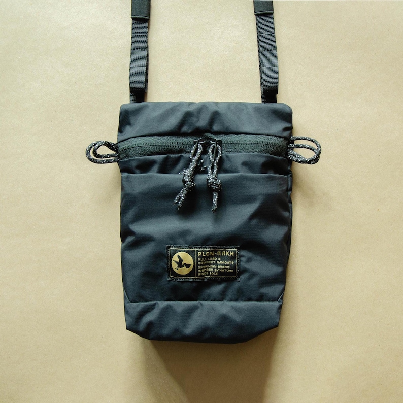 May include: A black crossbody bag with a zipper closure and a small patch with the text "PLCN-ПЛАЙК" and a bird logo. The bag has a black adjustable strap and a drawstring closure.