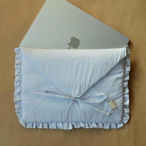 May include: A light blue and white gingham laptop sleeve with a ruffled edge and a bow closure. A silver laptop is partially visible inside the sleeve. The sleeve has a small tag attached.