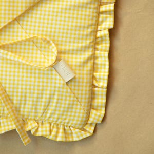 May include: A yellow and white gingham fabric item featuring a ruffled edge and a small, rectangular beige tag with the text PLCNY printed in black. The textile has a consistent checkered pattern throughout with a soft, folded appearance.