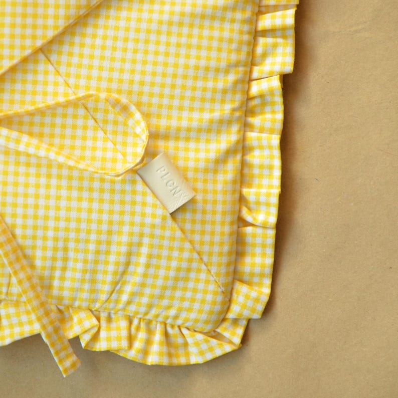 May include: A yellow and white gingham fabric item featuring a ruffled edge and a small, rectangular beige tag with the text PLCNY printed in black. The textile has a consistent checkered pattern throughout with a soft, folded appearance.