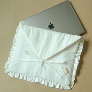 Può includere: Una custodia bianca per laptop con bordo a balze e chiusura con fiocco. La custodia è realizzata in tessuto morbido ed è progettata per un laptop. Un laptop argentato con il logo Apple è parzialmente visibile.