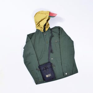 May include: A green utility jacket with a yellow hooded liner and a black crossbody bag. The jacket has a button closure and a flap pocket on the chest. A red and green baseball cap is also included.