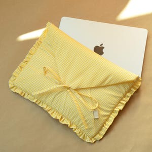 May include: A yellow and white gingham laptop sleeve featuring a ruffled edge and a fabric tie closure. The padded envelope-style case is constructed from checkered fabric and contains a silver laptop with a visible apple logo on the lid.