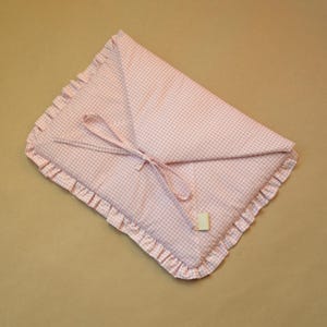 May include: A rectangular fabric pouch featuring a light pink and white gingham check pattern. The pouch has a ruffled edge trim and a triangular flap closure secured with two thin fabric ties. A small rectangular beige tag is attached to one tie.