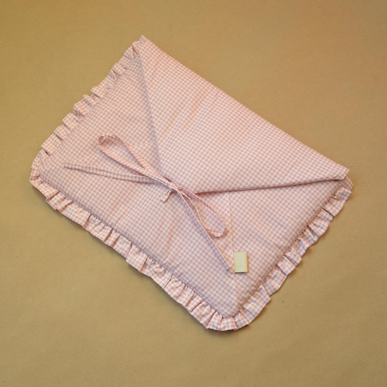 May include: A rectangular fabric pouch featuring a light pink and white gingham check pattern. The pouch has a ruffled edge trim and a triangular flap closure secured with two thin fabric ties. A small rectangular beige tag is attached to one tie.