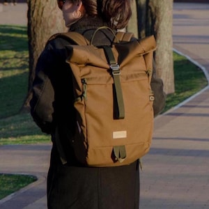 Peut inclure: Un sac à dos marron avec une fermeture à enroulement et des sangles vertes. Le sac à dos a une forme rectangulaire avec un petit patch rectangulaire sur le devant. Le sac est fait d'un matériau durable.